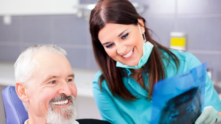 Dental professional in blue uniform smiles while assisting senior male patient in clinical office setting.