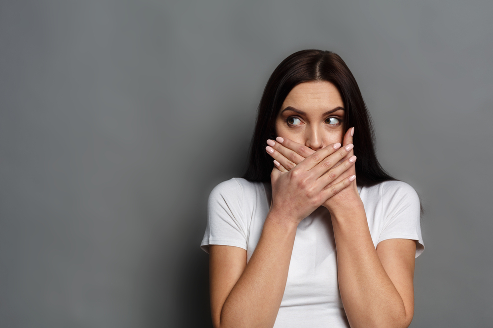 Woman in white shirt covering mouth with both hands against gray background, expressing surprise or concern.
