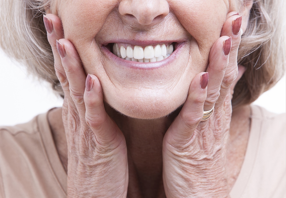 Smiling older woman touching her face and neck, displaying healthy skin and dental results.