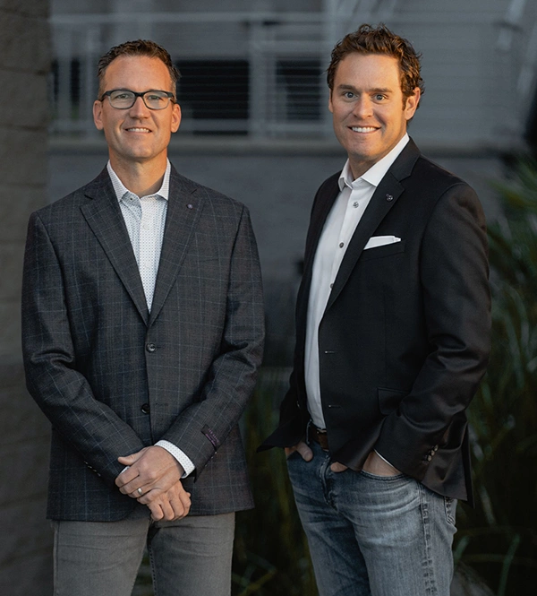 Two professional men in business casual attire posing together in a modern office setting.