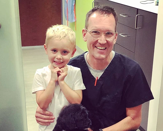 Adult male dentist with young blonde child and black dog in clinical office setting, smiling.