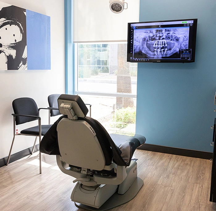 Modern dental operatory with treatment chair, mounted monitor displaying dental imaging, and waiting area seating.
