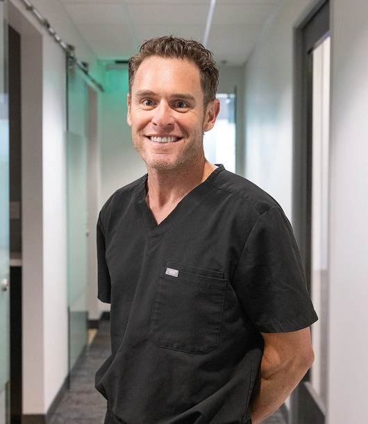 Male dentist or oral surgeon in black scrubs smiling in modern dental office hallway.